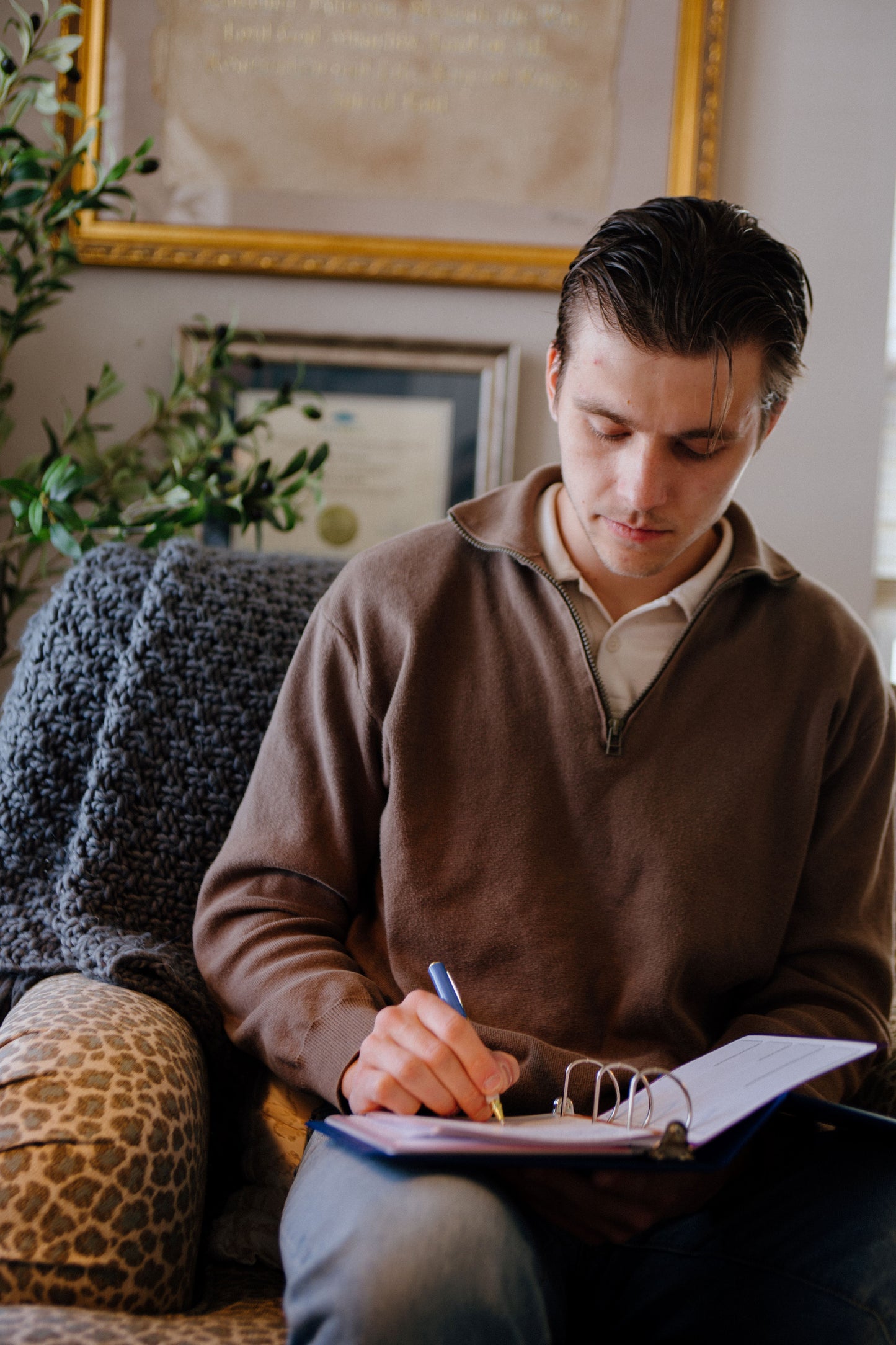 Man sitting on a couch writing in a notebook with a cozy indoor setting.